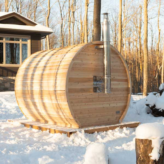 Wooden barrel-shaped sauna in a snowy outdoor setting with a cabin in the background.